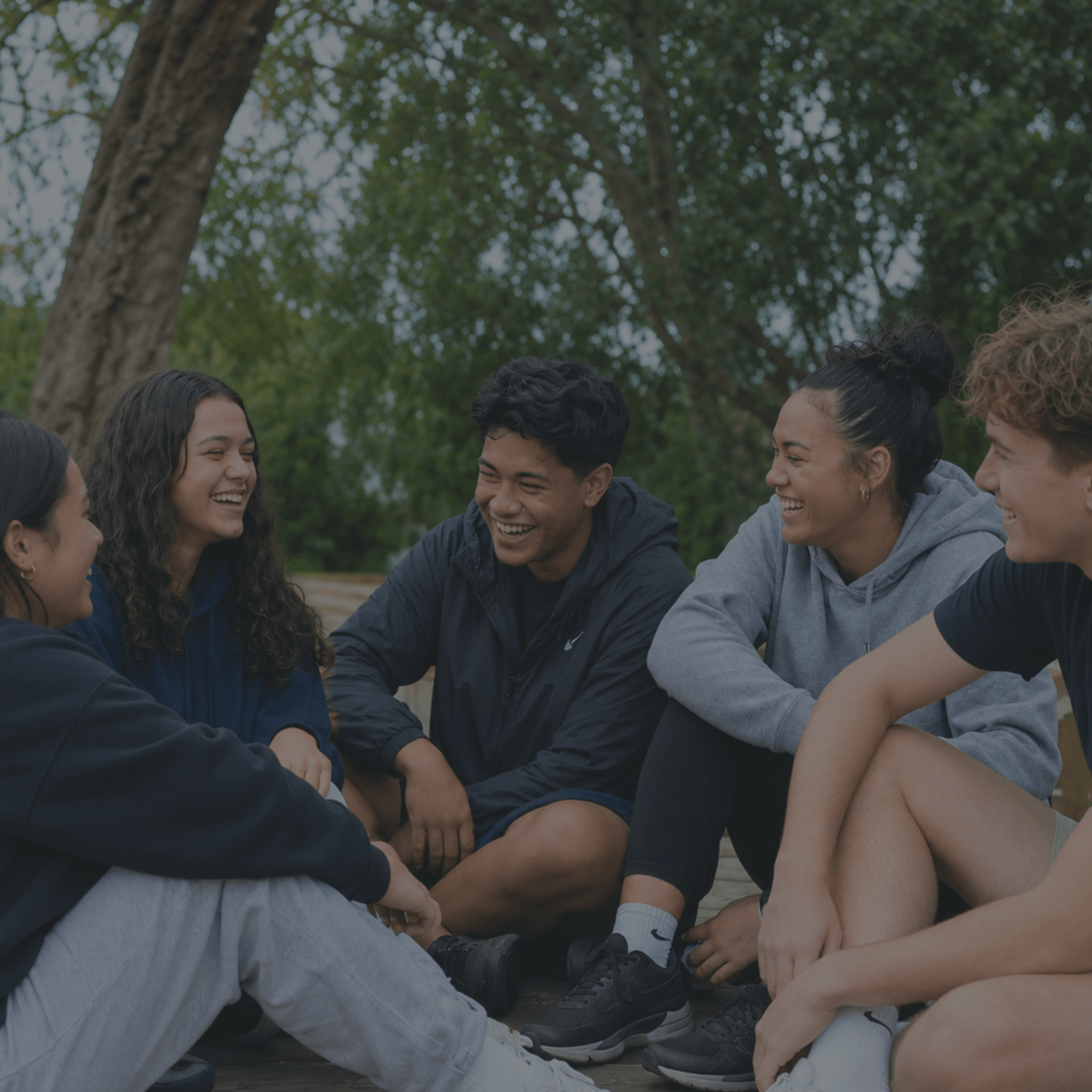 pacific-maori-pakeha-youth-group-outdoors-circle-laughing-dark-overlay-nz